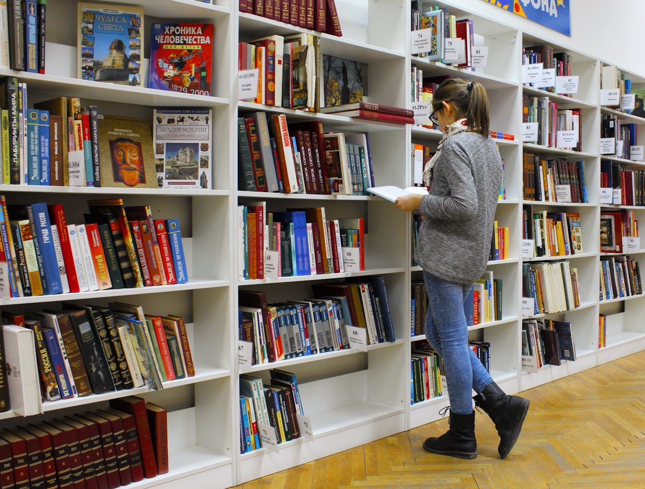 services-04 A young woman stands reading a book in a well-stocked library.